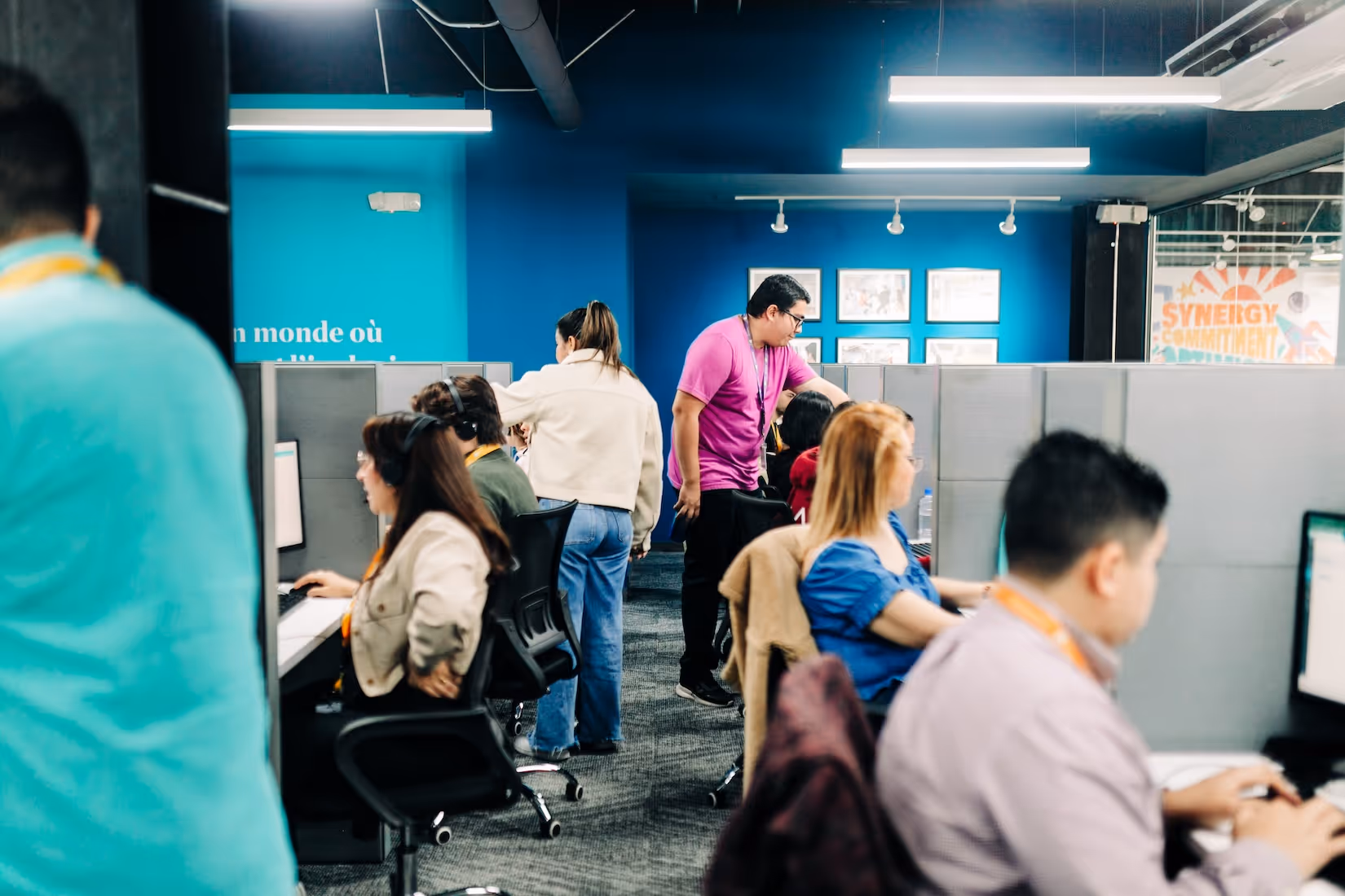Busy office with employees working at computer cubicles, some wearing headsets, and one person standing and assisting another.