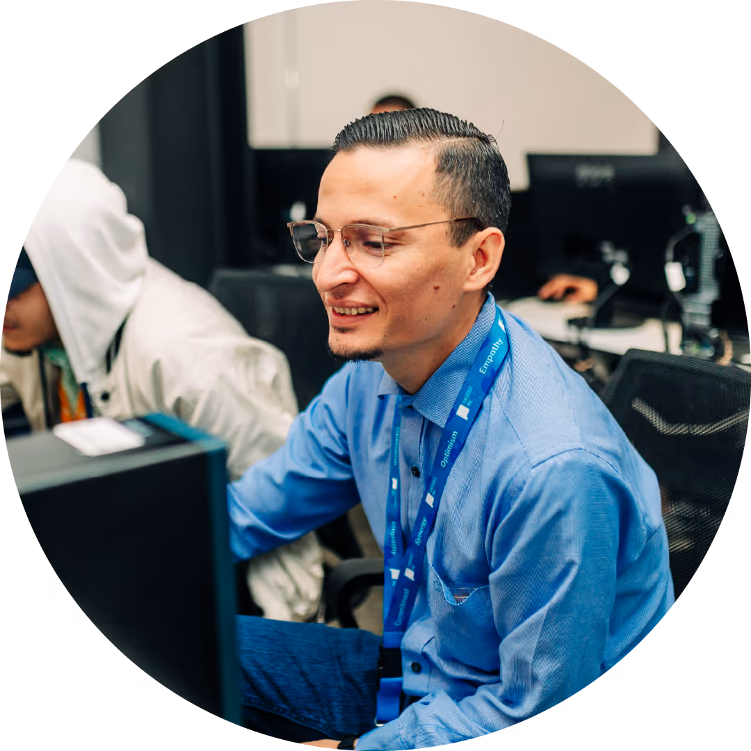 Smiling man wearing glasses and a blue shirt sitting at a computer workstation with a blue lanyard around his neck.