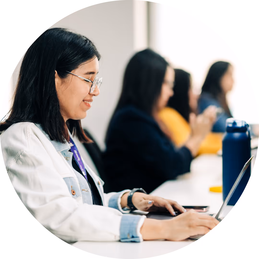 Woman wearing glasses and a white jacket working on a laptop at a desk, with other people blurred in the background.