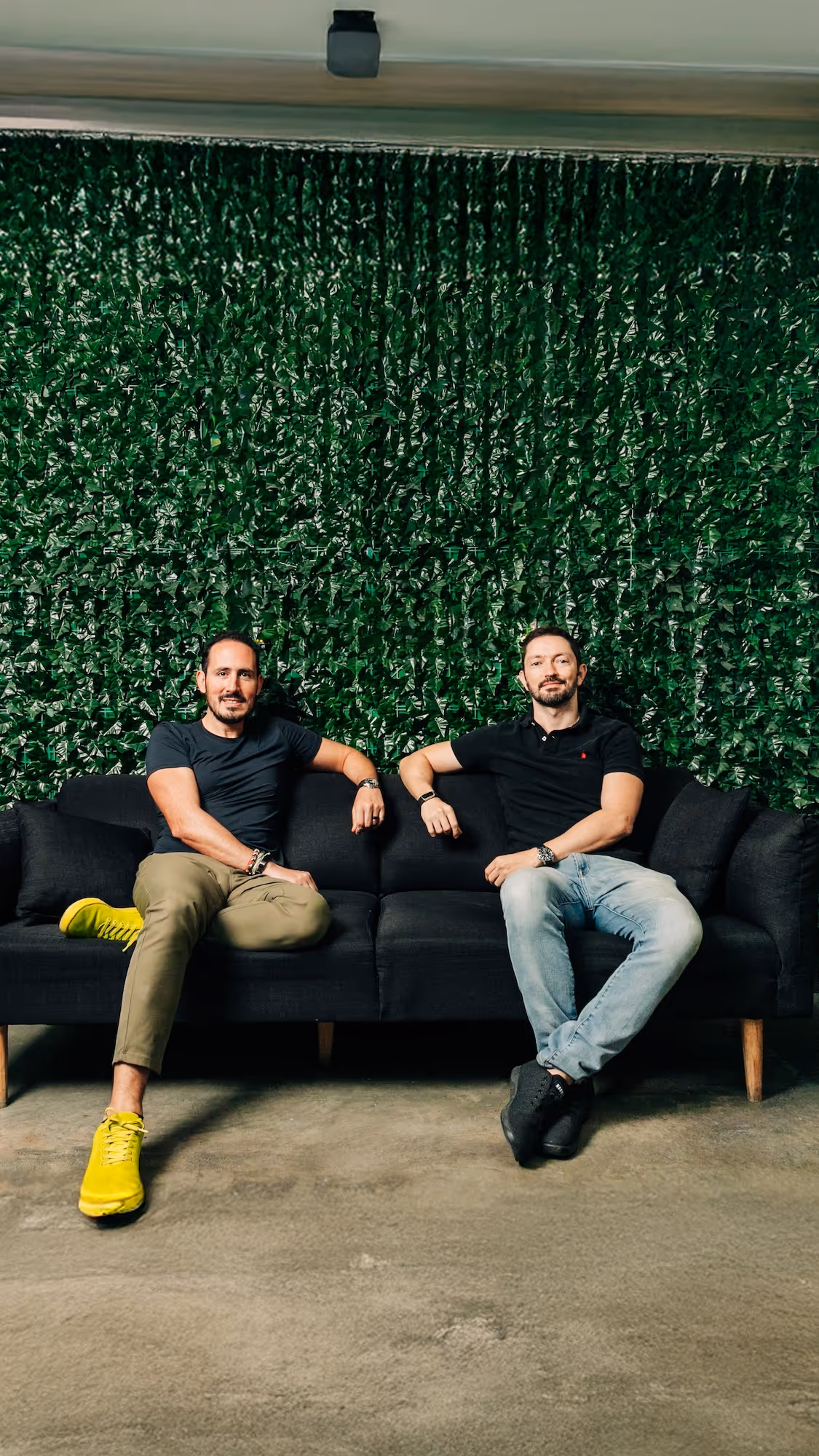 Two men sitting on a black couch in front of a green leafy wall, one wearing yellow shoes and the other wearing black shoes.