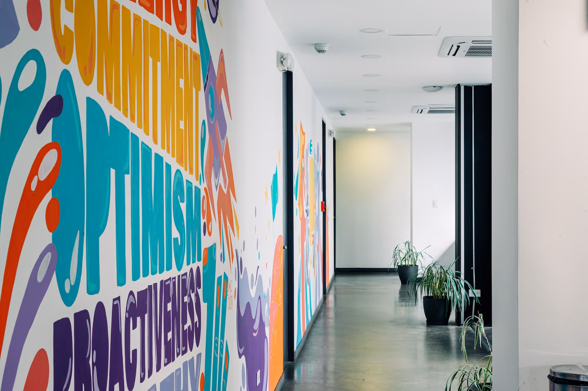 Bright hallway with colorful motivational words including optimism and proactiveness painted on the left wall and potted plants on the right side.