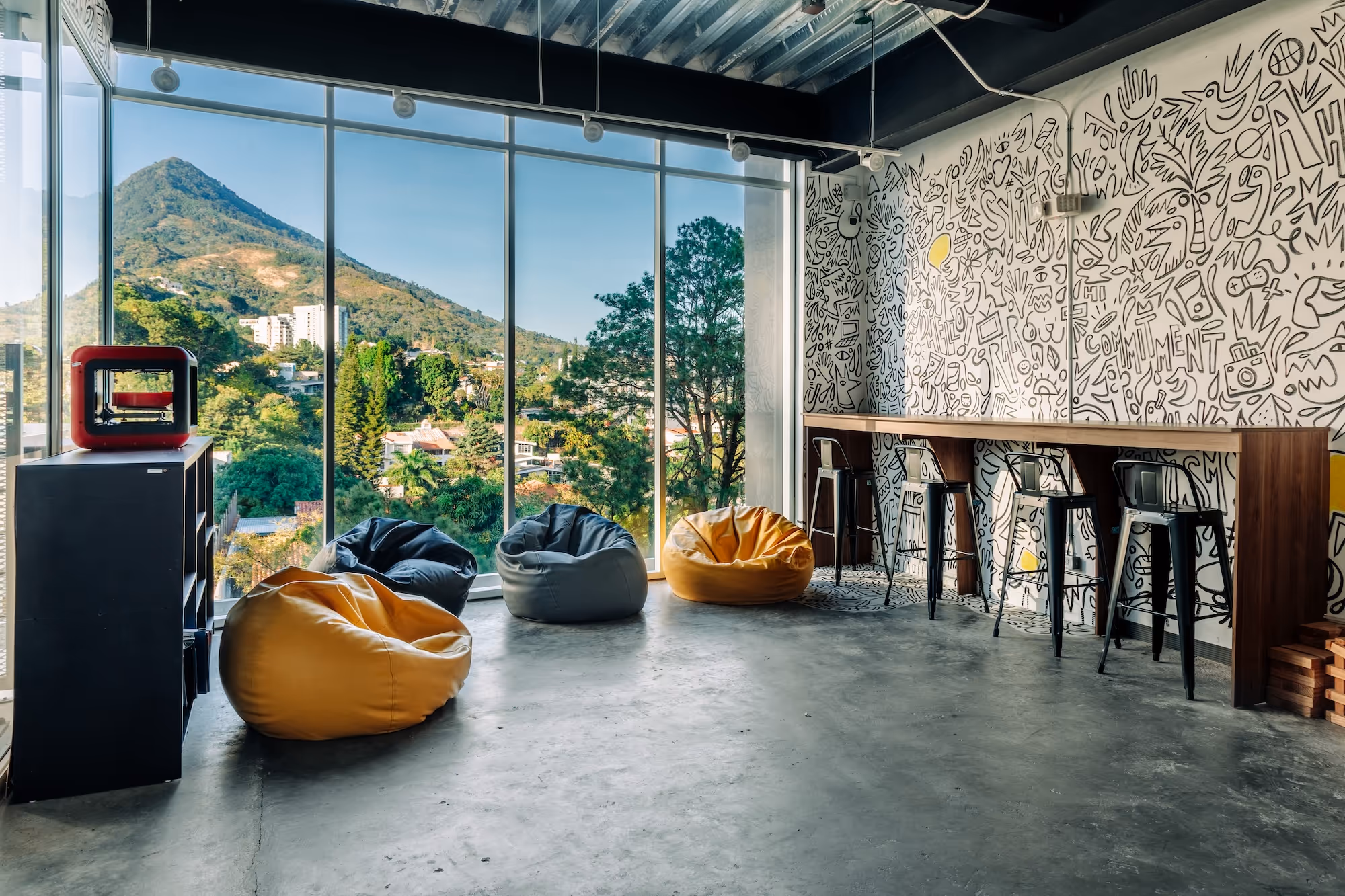 Modern lounge area with floor-to-ceiling windows showing a mountain view, colorful bean bags, bar stools, and a wall covered in black and white doodles.