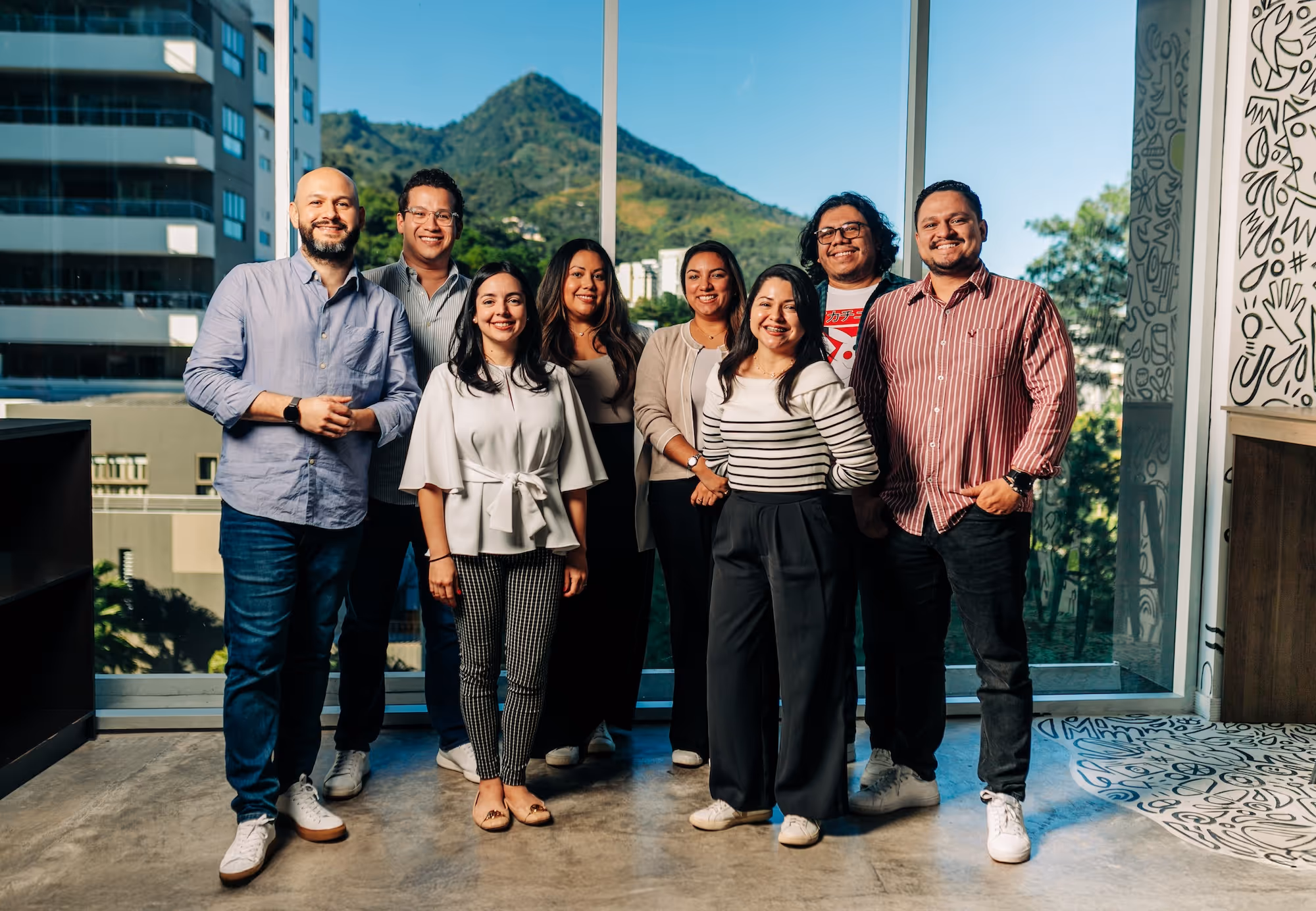 Group of seven smiling people standing indoors in front of large windows with a mountain and buildings visible outside.