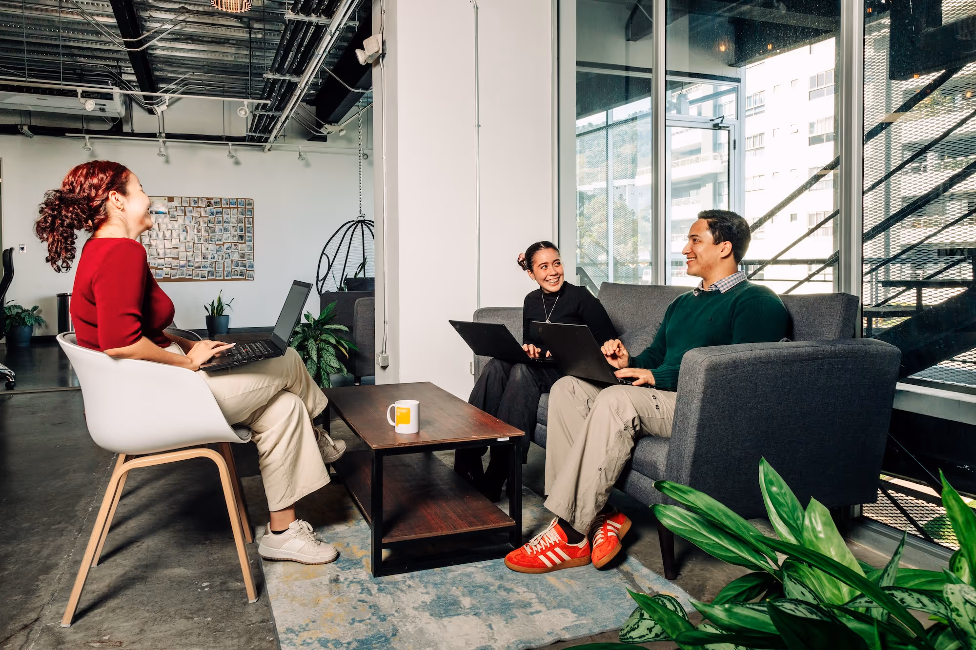 Three people in a modern office sitting on chairs and a sofa, smiling and working on laptops around a wooden coffee table with a mug.