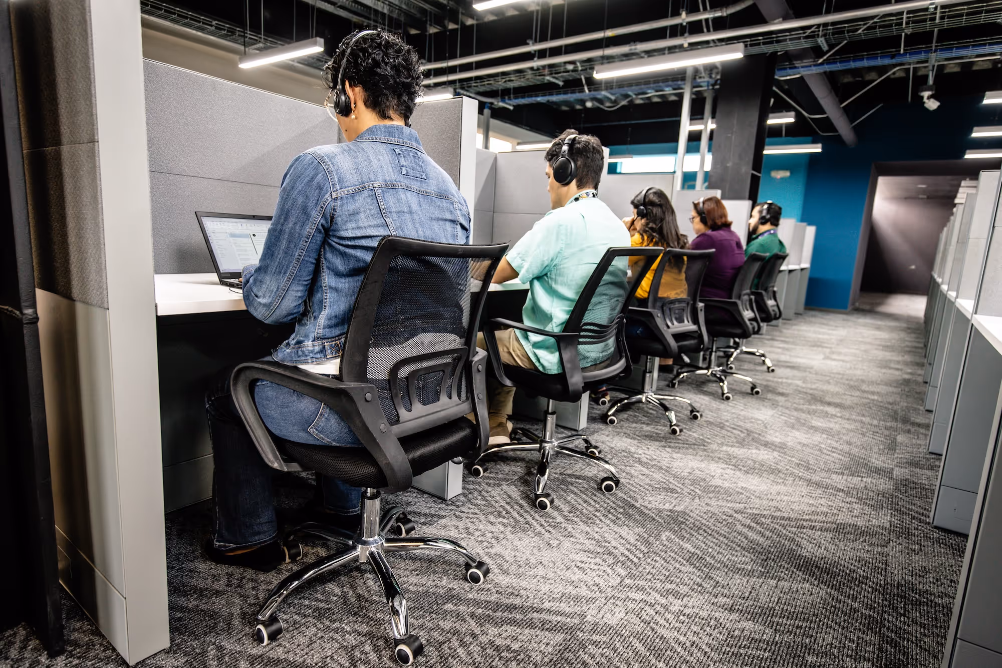 Five people wearing headsets seated at individual cubicles working on computers in a modern office.