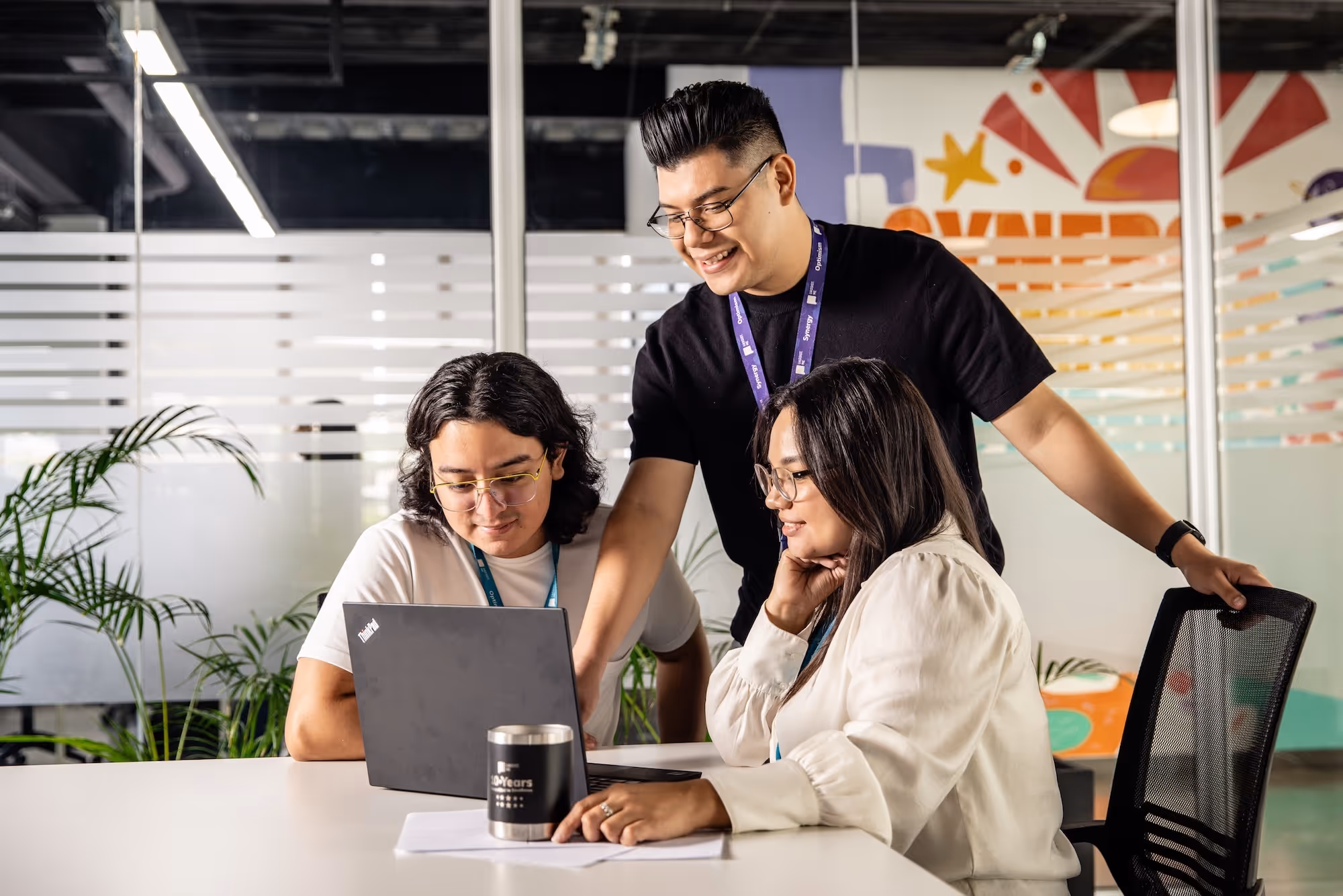 Three colleagues collaborating around a laptop in a modern office with indoor plants and glass partitions.