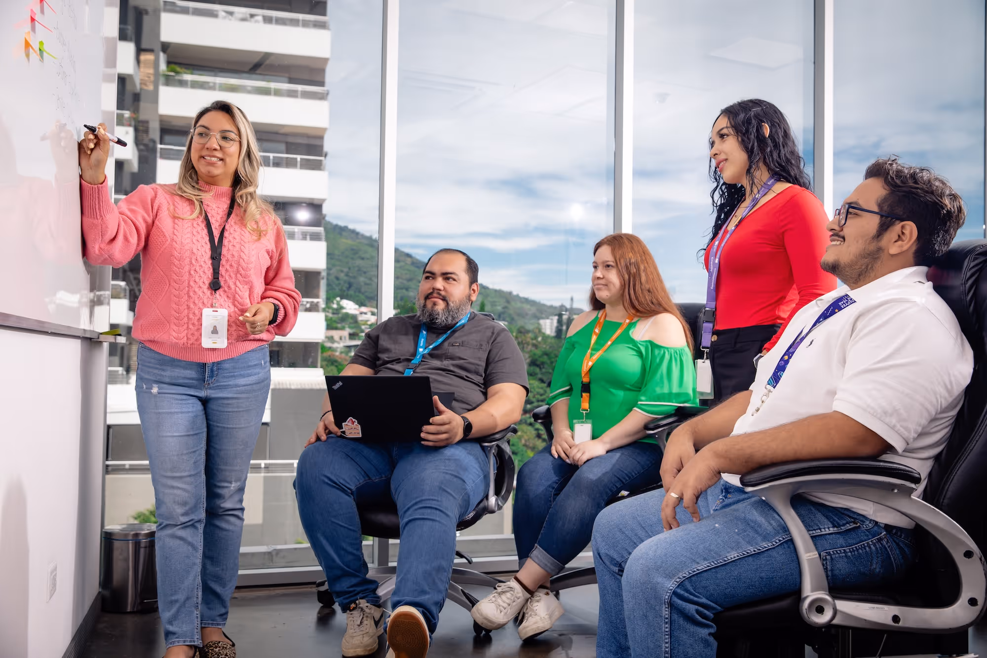 Woman in pink sweater writing on a whiteboard while four colleagues seated and standing watch attentively in a modern office with large windows.