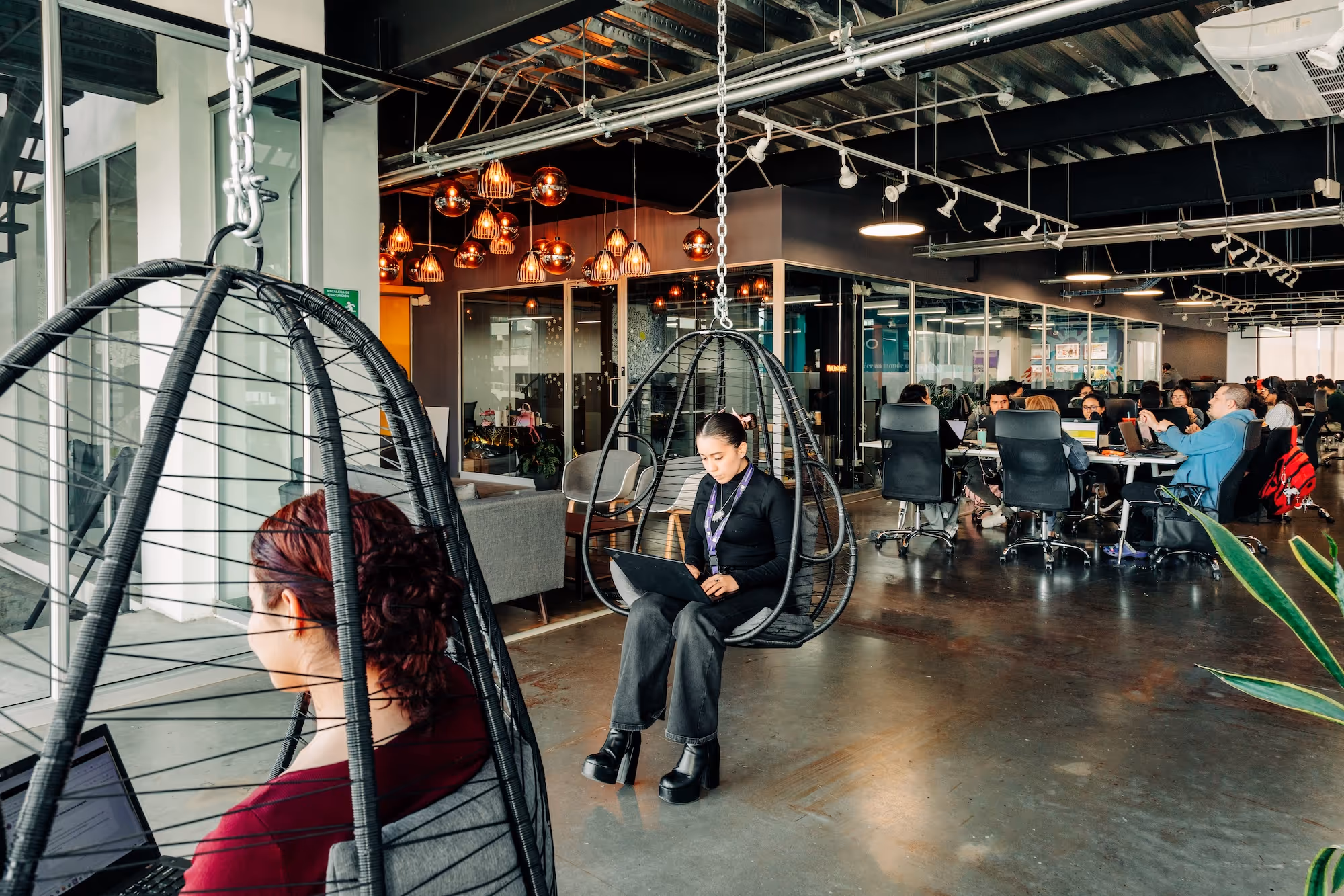 Two women working on laptops while sitting in hanging egg chairs inside a modern open office space with other people working at desks in the background.