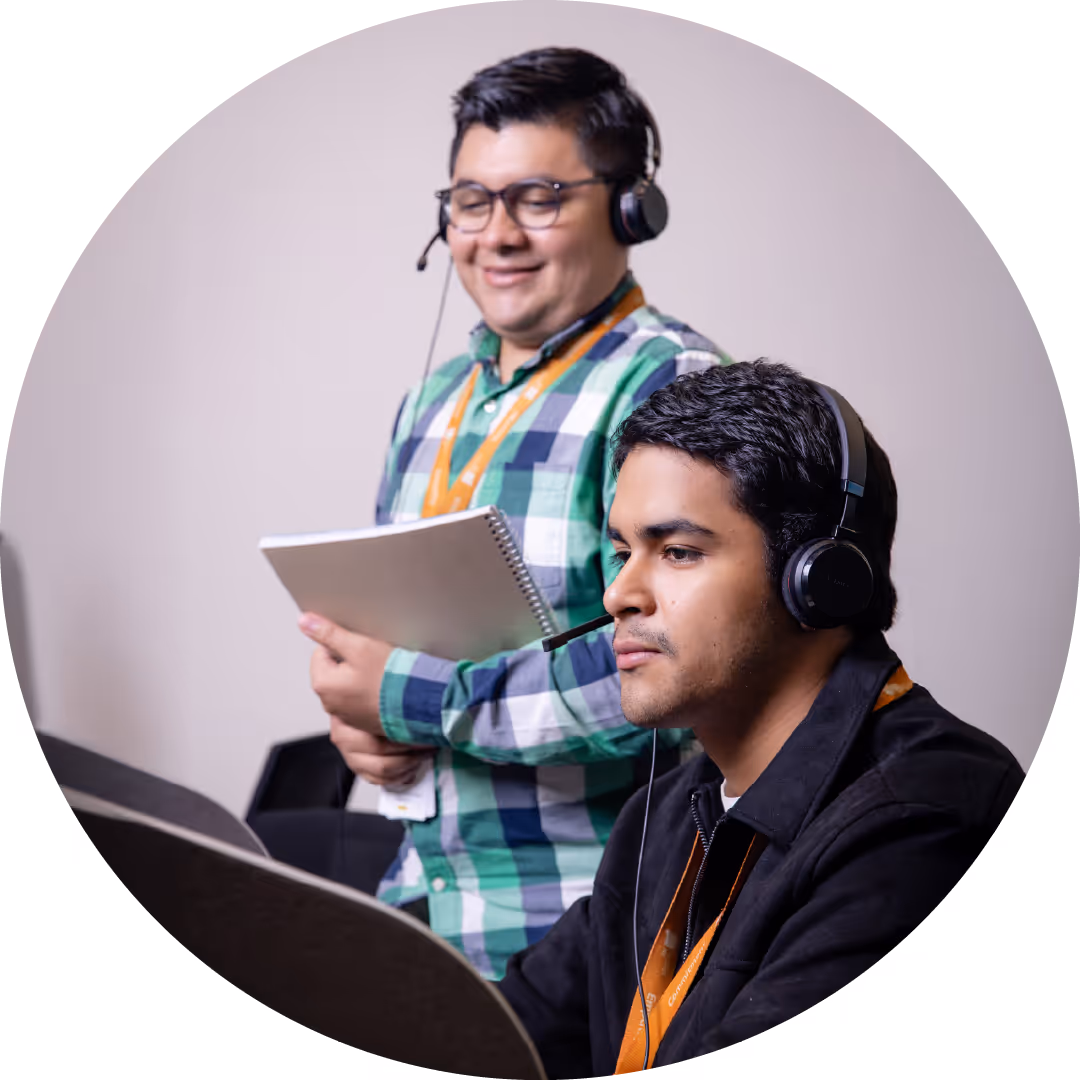 Two men wearing headsets working together, one seated focused on a computer, the other standing holding a notebook and smiling.