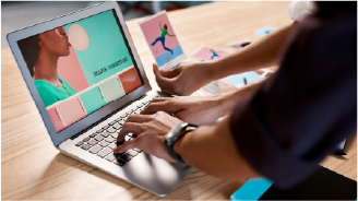Man with tattoos wearing a headset and plaid shirt working on a computer in an open office space.
