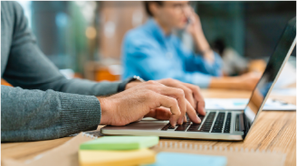 Man with tattoos wearing a headset and plaid shirt working on a computer in an open office space.