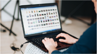 Man with tattoos wearing a headset and plaid shirt working on a computer in an open office space.