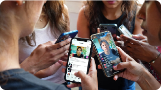 Man with tattoos wearing a headset and plaid shirt working on a computer in an open office space.