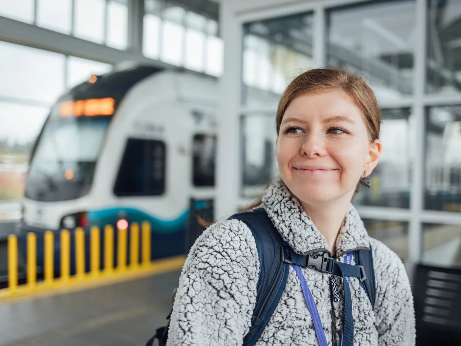 Young woman smiling at a station with a light rail train in the background.