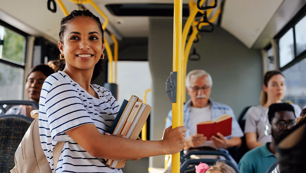 A woman smiling while riding a bus, wearing wireless earbuds and holding her phone, with other passengers seated around her.