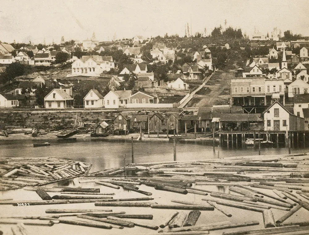 View of Seattle in 1880 from Yesler Wharf, showing houses on the hillside and logs floating in the harbor awaiting milling.