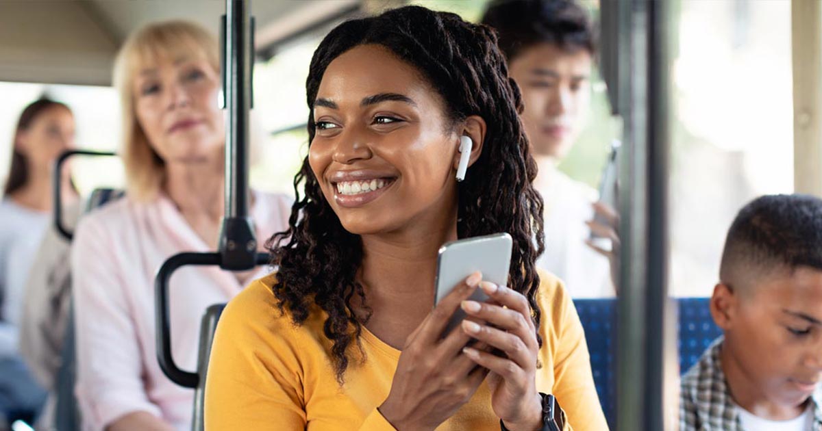 A woman smiling while riding a bus, wearing wireless earbuds and holding her phone, with other passengers seated around her.