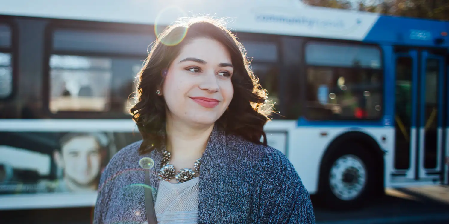 A woman smiling while riding a bus, wearing wireless earbuds and holding her phone, with other passengers seated around her.