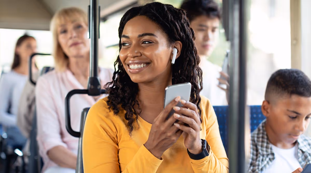 A woman smiling while riding a bus, wearing wireless earbuds and holding her phone, with other passengers seated around her.