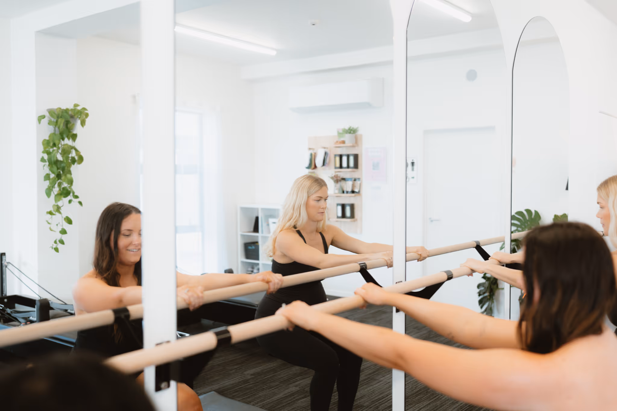 Women holding a barre and doing squats in a bright fitness studio with mirrors and plants.