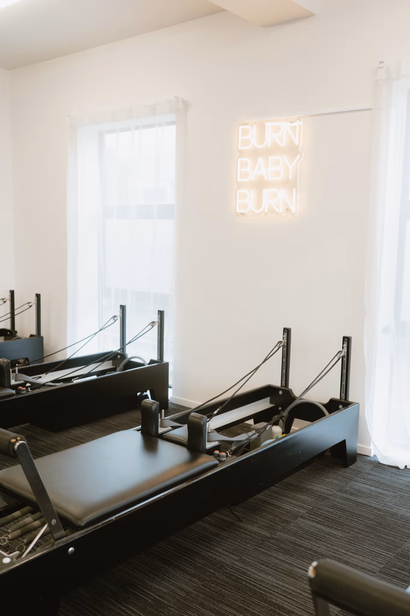 Modern Pilates studio with black reformer machines and a neon sign saying Burn Baby Burn on white wall.