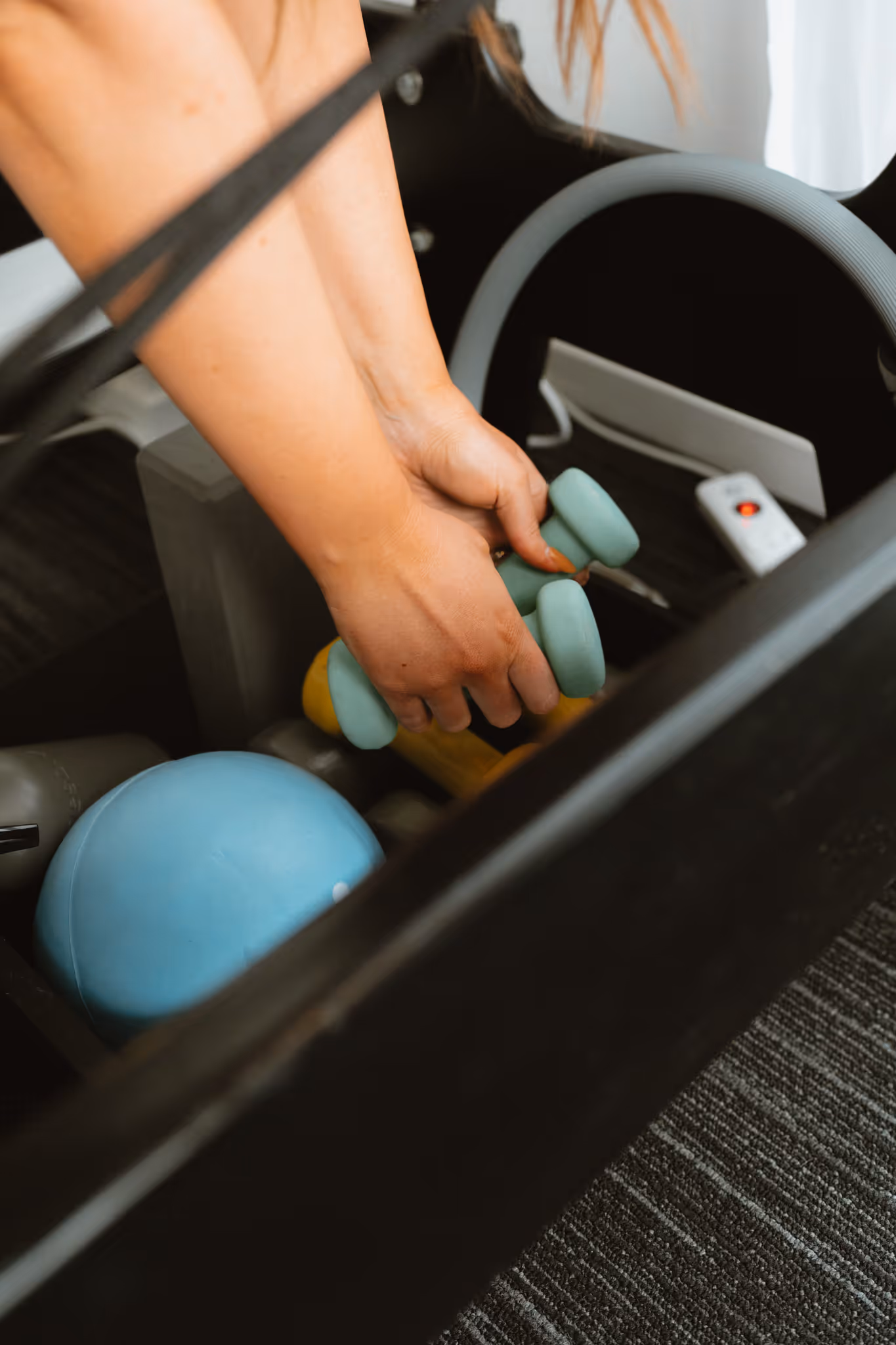 Hands holding light blue dumbbells over a container with various exercise equipment inside.