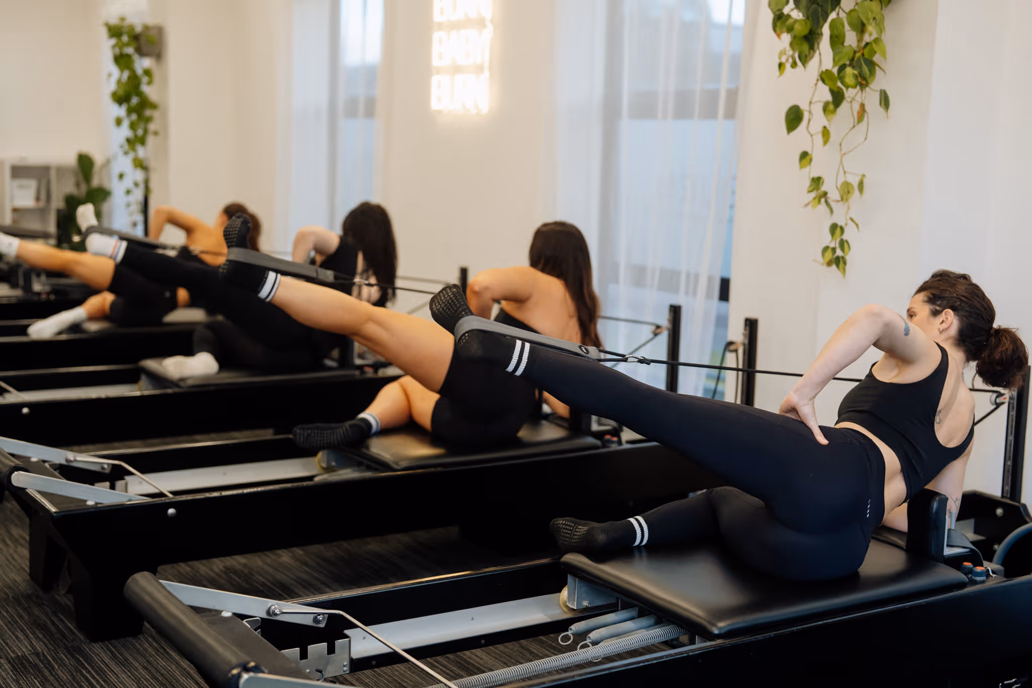 Four women in black workout clothes doing leg exercises on Pilates reformer machines in a bright studio.
