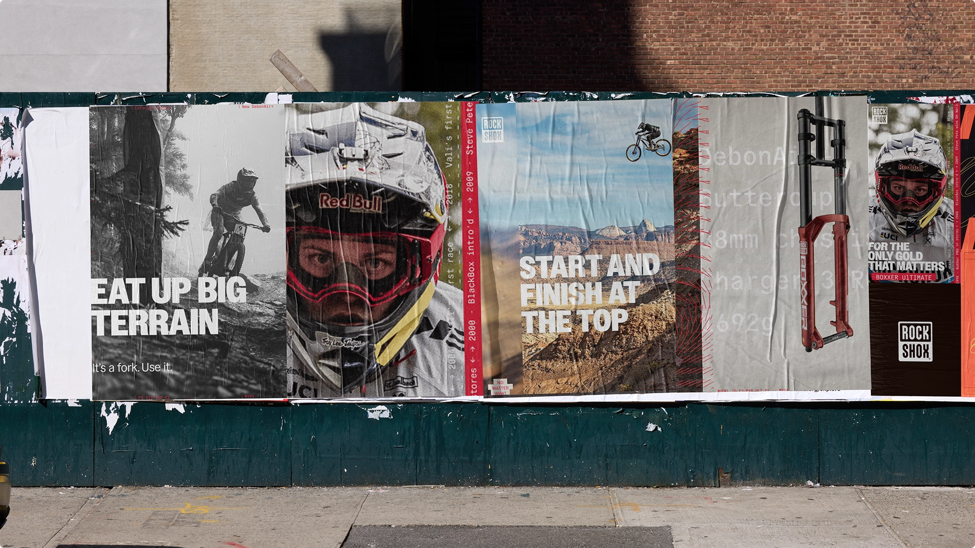 A series of outdoor posters on a wall featuring mountain biking: a black and white photo of a biker on rough terrain with 'EAT UP BIG TERRAIN' text, a close-up of a helmeted Red Bull rider, a rider jumping over a canyon with 'START AND FINISH AT THE TOP' text, a bike suspension fork image, and a Red Bull biker in full gear with 'FOR THE ONLY GOLD THAT MATTERS' text.