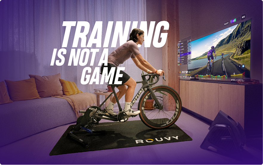 Person cycling on a stationary ROUVY bike facing a TV screen displaying a virtual cycling route, with text saying 'TRAINING IS NOT A GAME'.