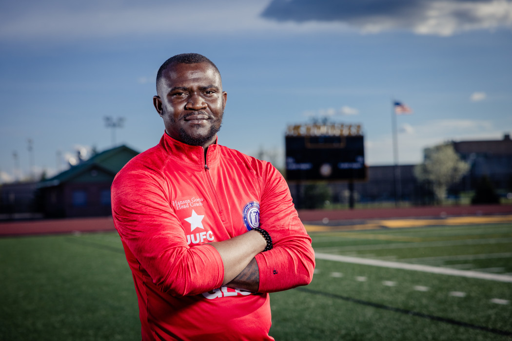 Man in a red sports jacket standing on a football field with arms crossed, with an American flag and scoreboard in the background.