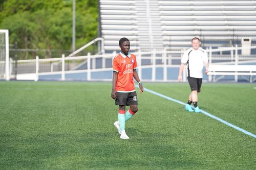 Soccer player in an orange jersey and black shorts walking on a green field with empty bleachers in the background.