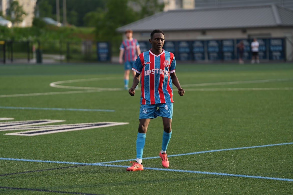 Young soccer player in a red and blue striped uniform standing on a green soccer field during a game.