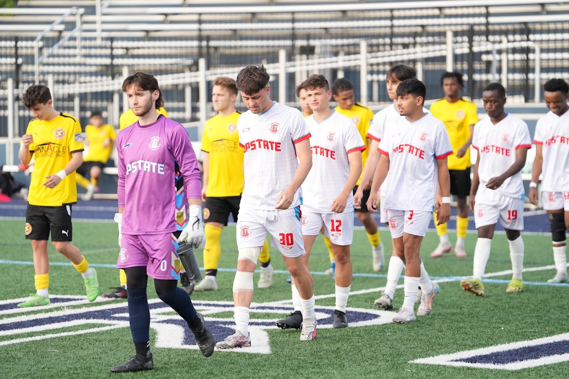 Two soccer teams walking on the field, one in white jerseys with red UPSTATE lettering and another in yellow jerseys.