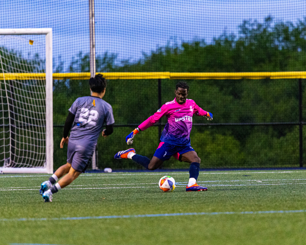 Soccer goalkeeper in pink jersey kicking the ball while an opponent in gray jersey approaches near the goal.