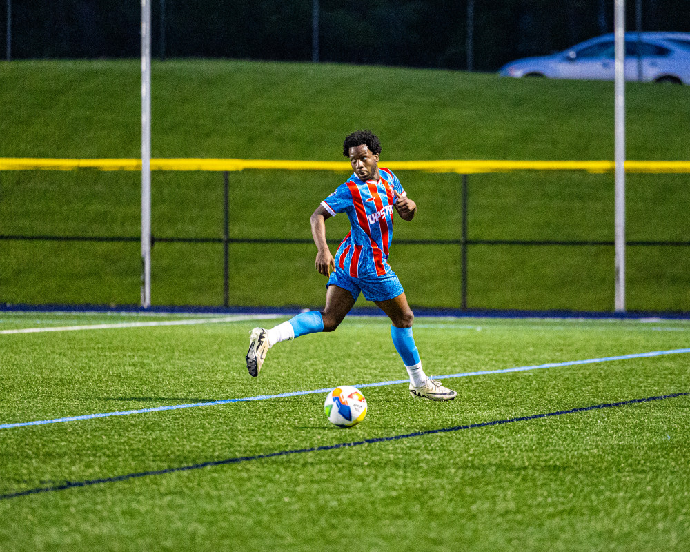 Soccer player in blue and red striped uniform kicking a colorful ball on a green field at night.