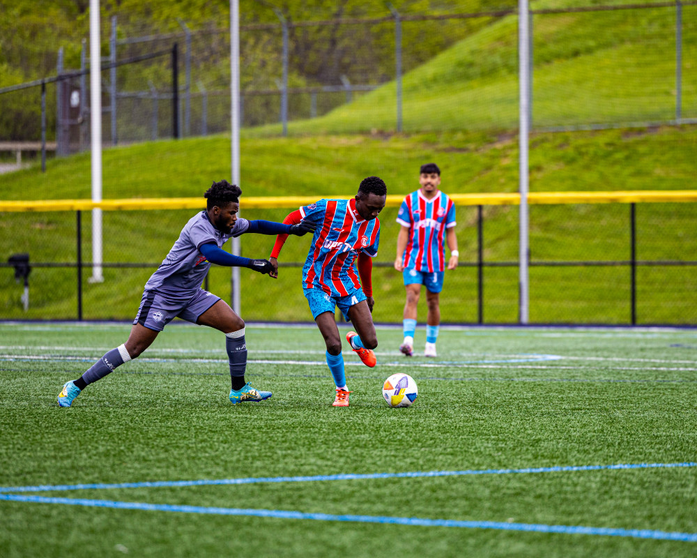 Two soccer players competing for the ball on a green field with one player in gray reaching to block a player in red and blue stripes, another player stands in the background.