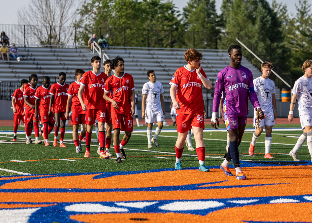 Soccer players from the Upstate team in red and white uniforms walking on a field with orange and blue markings.