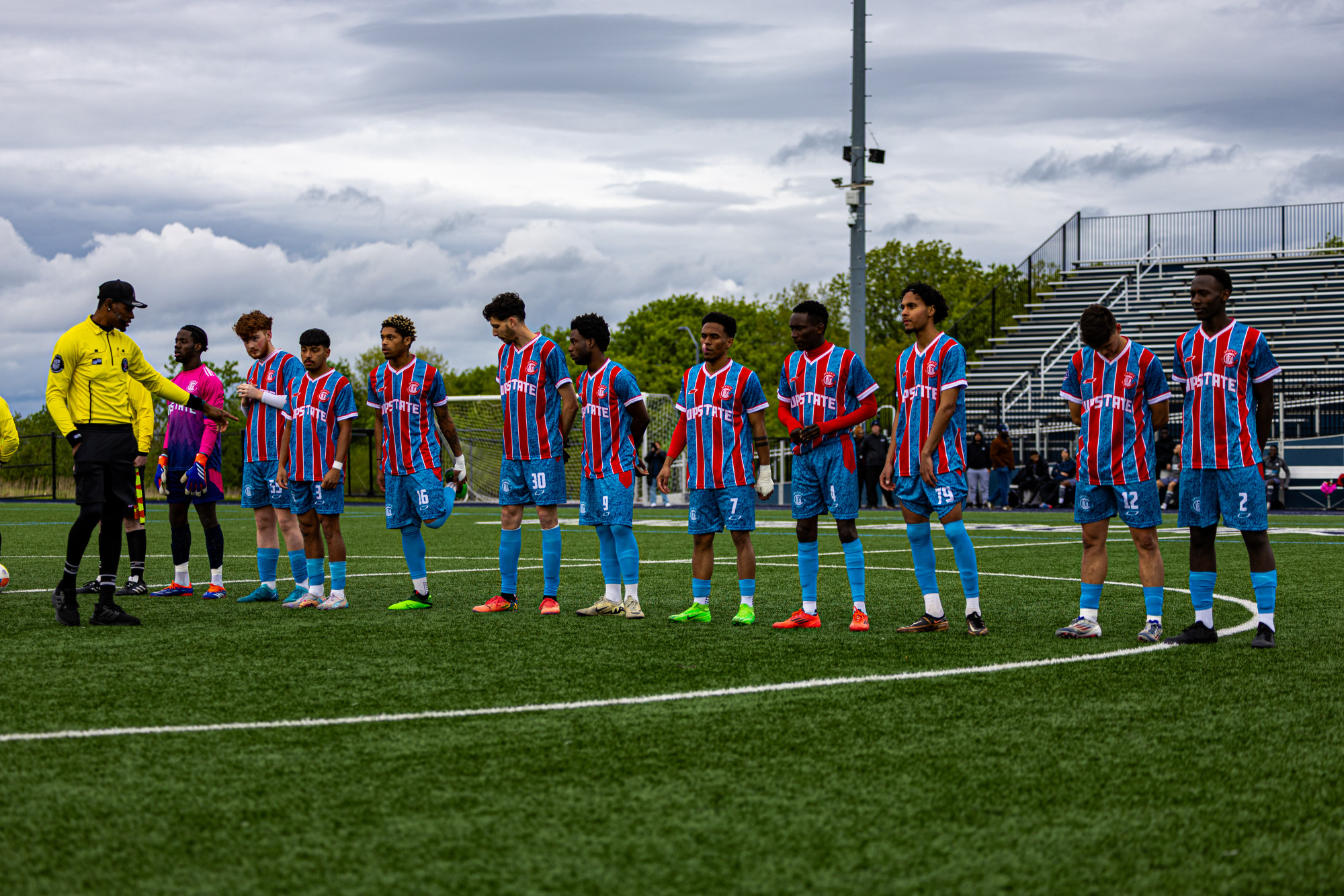 Soccer team lined up on the field wearing blue, red, and white striped jerseys with blue shorts, under a cloudy sky.