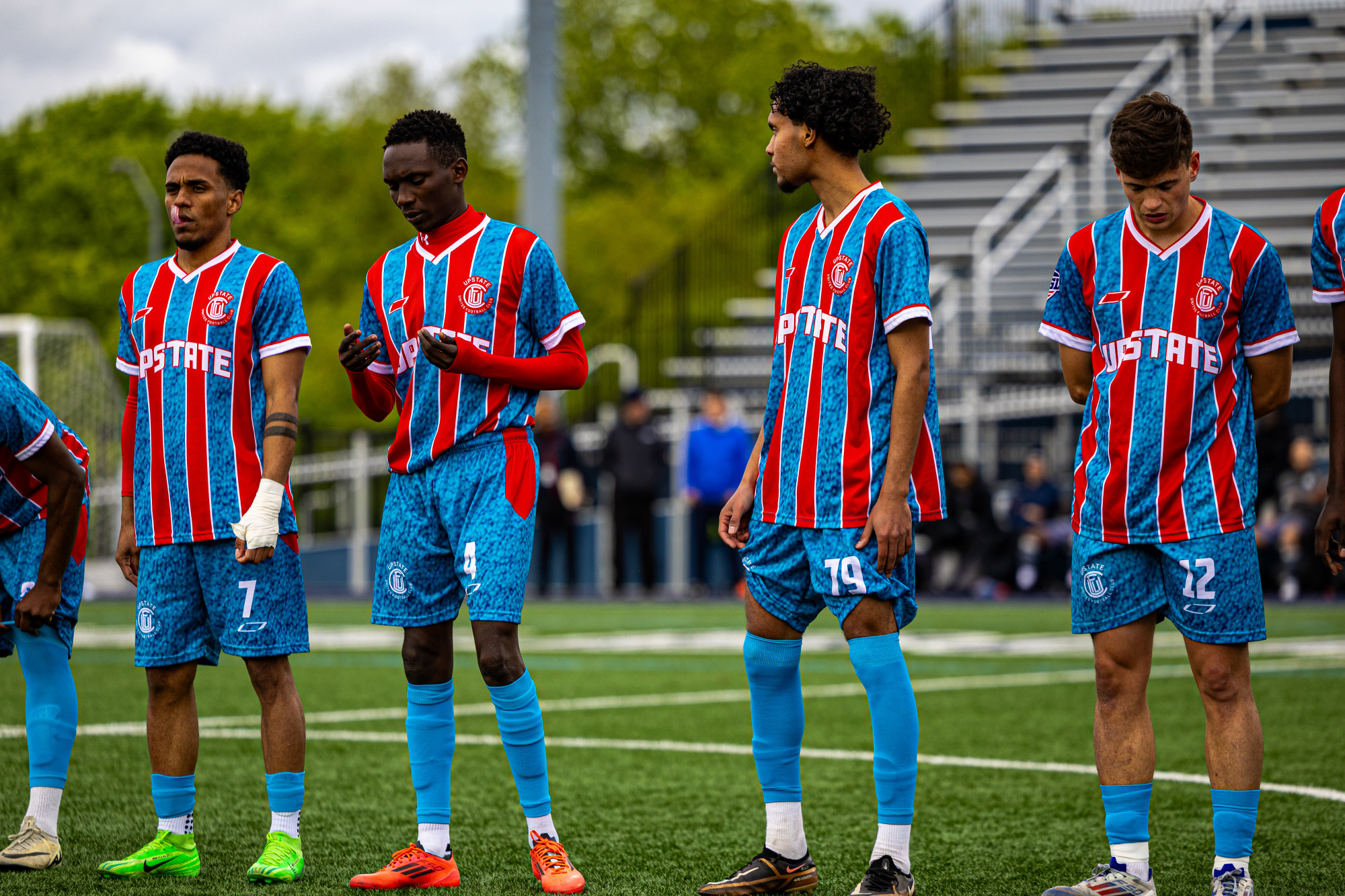 Four soccer players in red and blue striped Upstate uniforms standing on a field before a game.