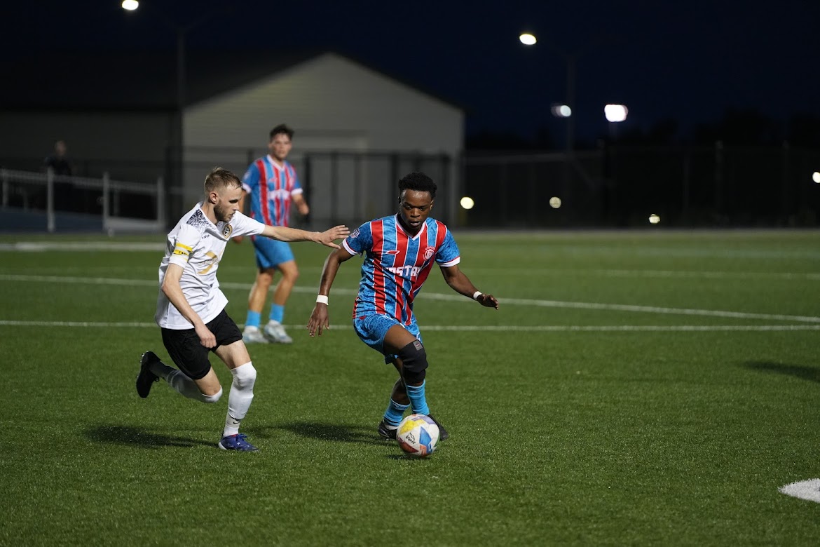 Soccer player in blue and red striped jersey dribbling the ball while a player in white tries to defend on a lit field at night.