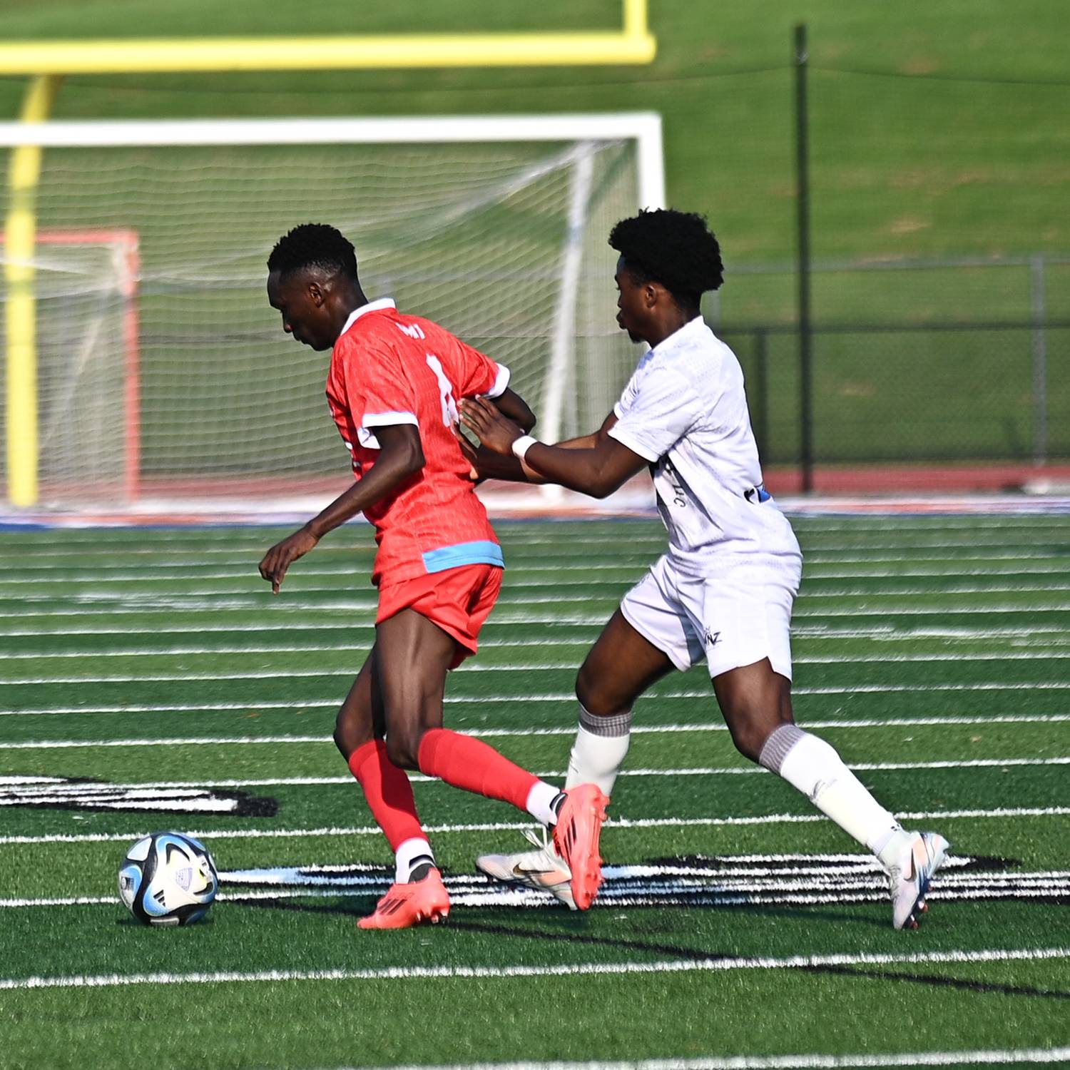 Two young male soccer players competing for the ball on a green turf field with goalposts in the background.