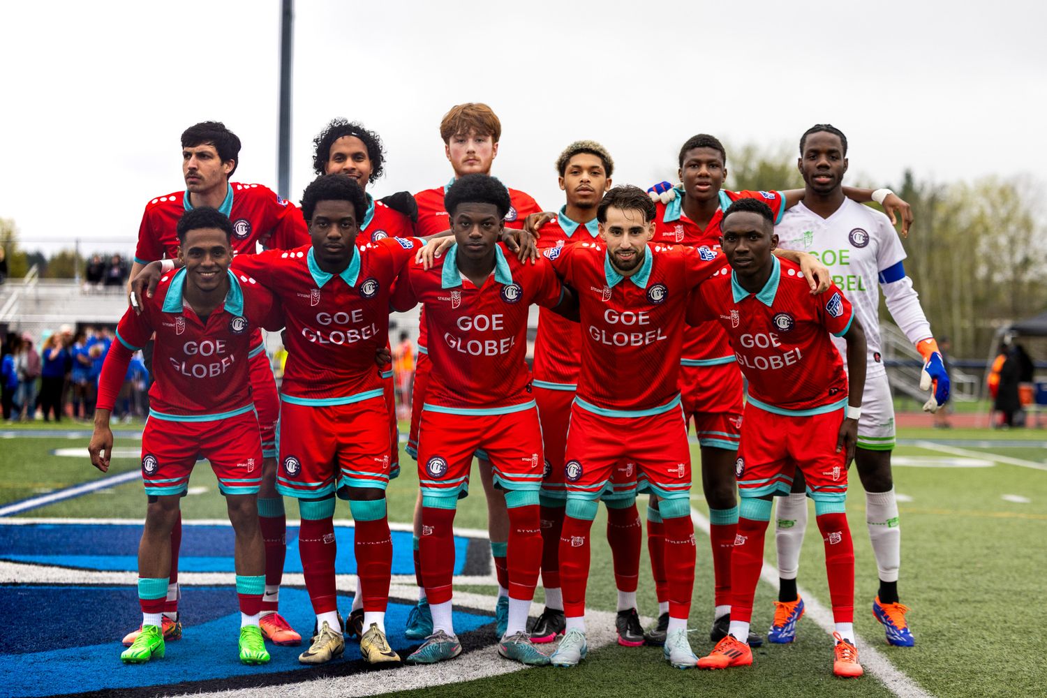 Soccer team in red uniforms with turquoise trim posing together on a field, with the goalkeeper in a white uniform.