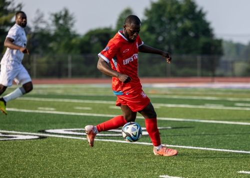Soccer player in red uniform controlling a soccer ball on a green field with another player in white running behind.