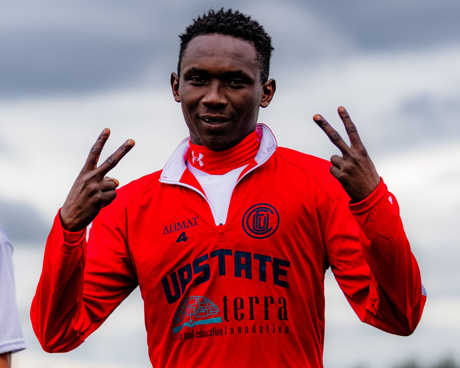 Man wearing a red sports jacket with 'UPSTATE' and other logos, holding up two peace signs with both hands.