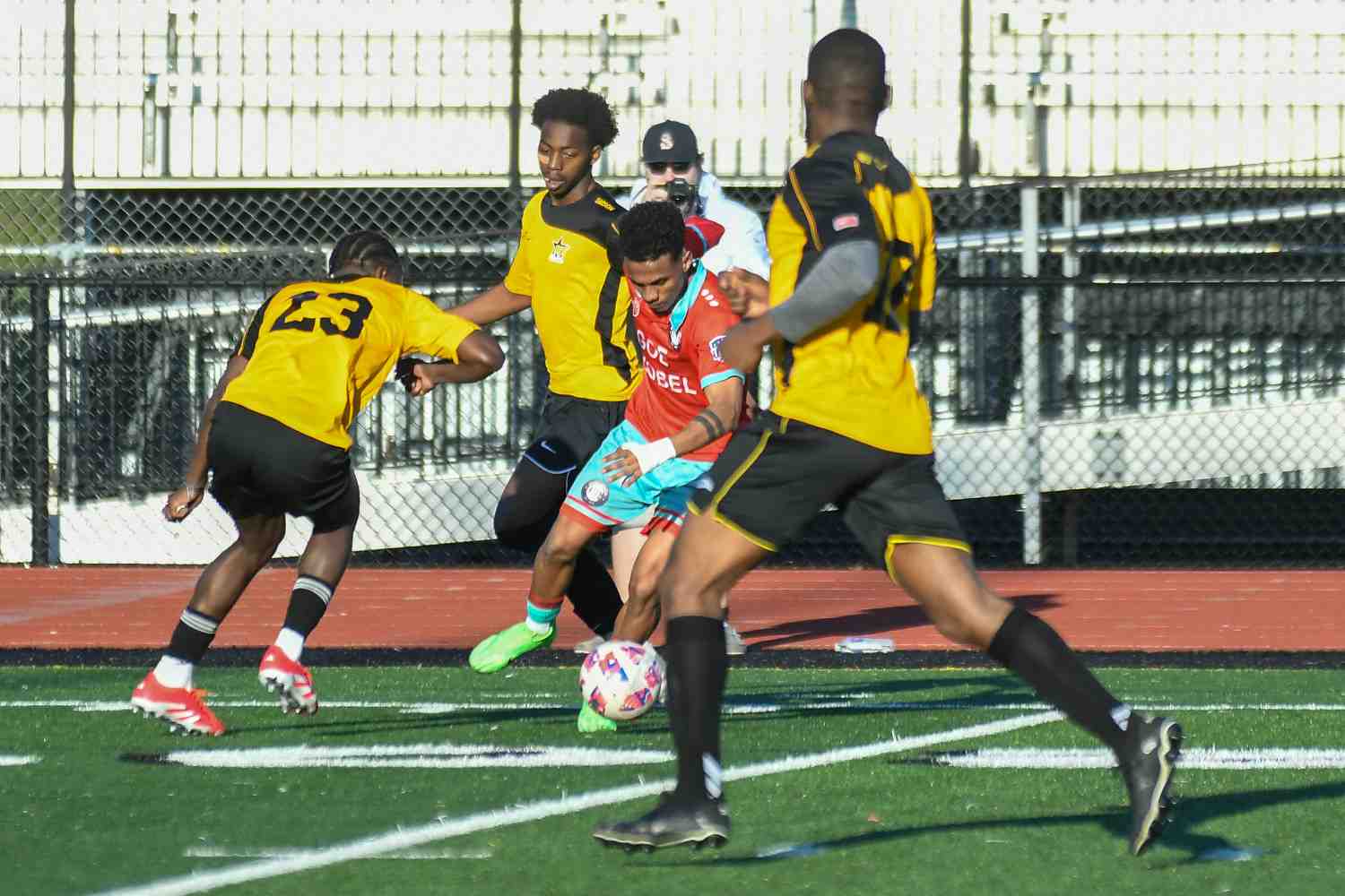 Soccer player in red and blue uniform dribbling the ball while three opponents in yellow and black uniforms try to defend.