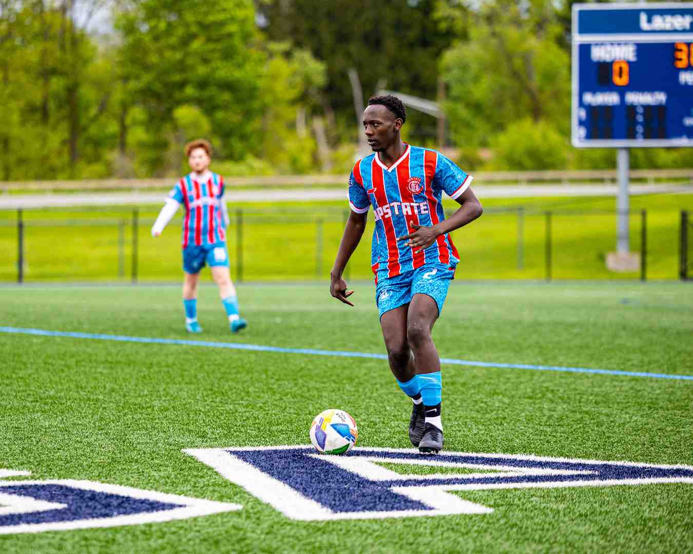 Soccer player in blue and red striped uniform dribbling a ball on a green field near the sideline, with a scoreboard and another player in the background.