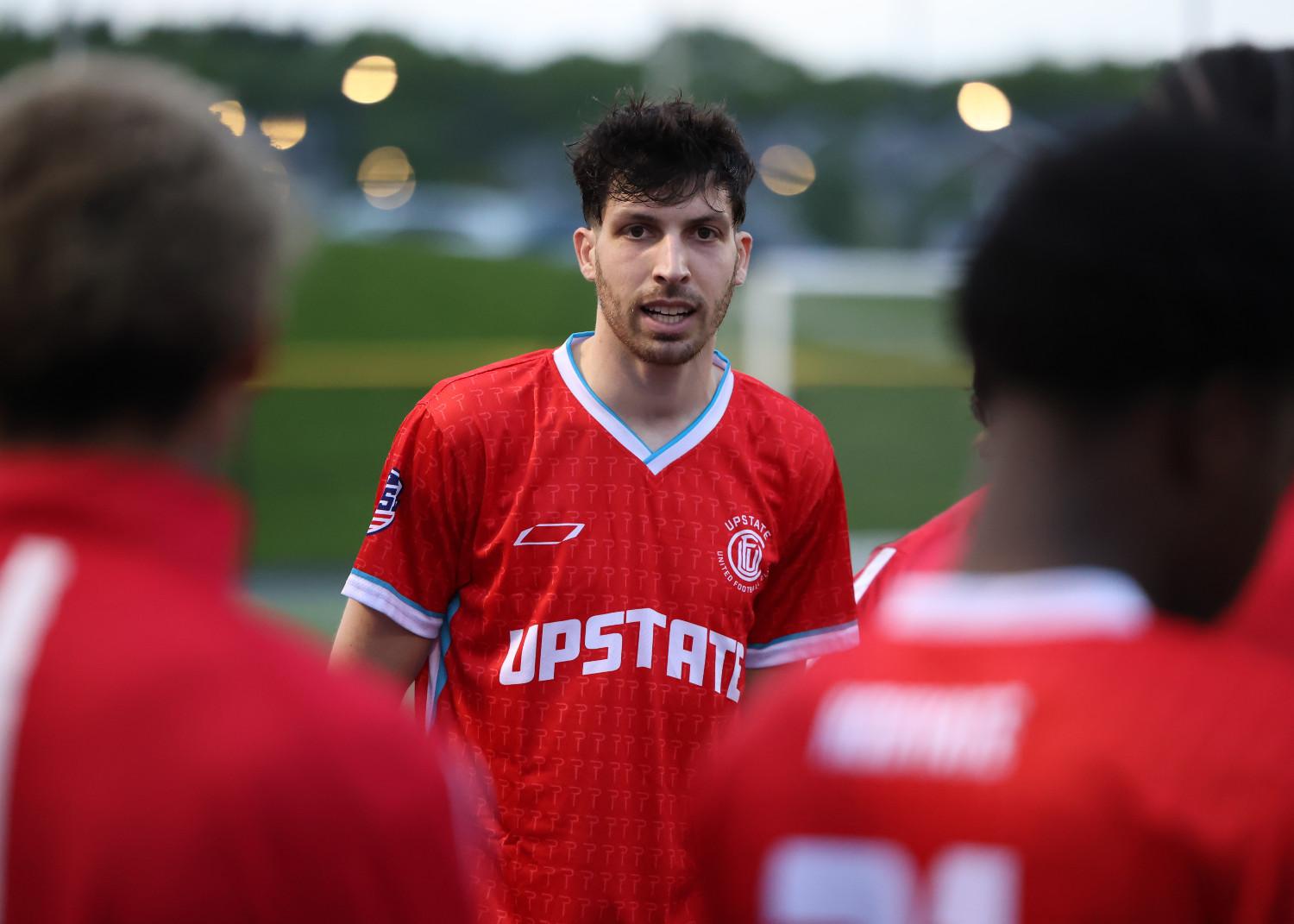Soccer player in red Upstate United Football Club jersey speaking to teammates on the field.