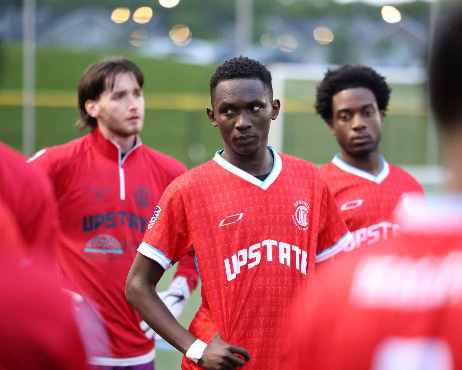 Three soccer players wearing red Upstate jerseys standing outdoors on a field, looking focused and thoughtful.