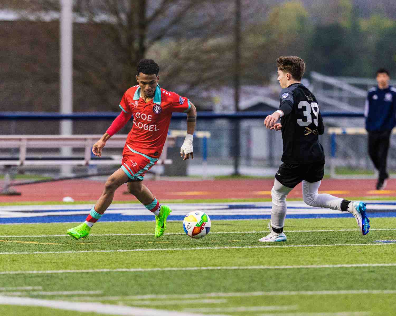 Soccer player in red jersey and neon green cleats preparing to kick the ball while an opponent in black jersey and white leggings approaches on the field.