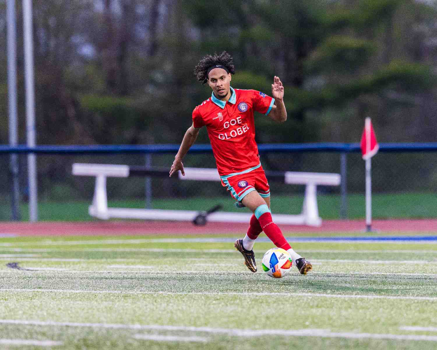 Soccer player wearing red and light blue uniform kicking a ball on a green field near a corner flag.